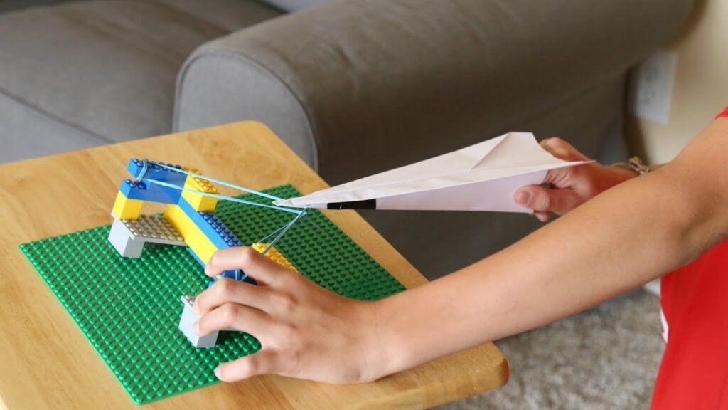 Child's hands operate a blue and yellow LEGO catapult on a green baseplate, launching a paper airplane on a wooden table.