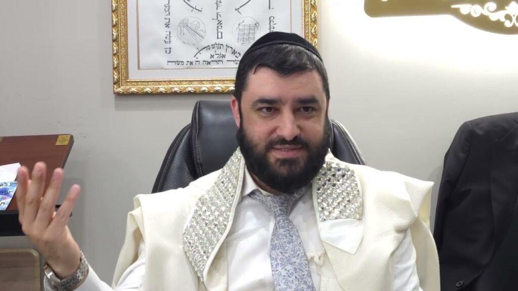 Bearded man wearing a white suit and kippah sits in an office, gesturing with his left hand.