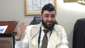 Bearded man wearing a white robe with silver embellishments and a kippah with a small camera mount, gesturing with his hand in an office setting with Hebrew framed artwork on the wall behind him.