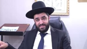 Man in a black hat and suit sits in an office, smiling at the camera with a framed document on the wall behind him.