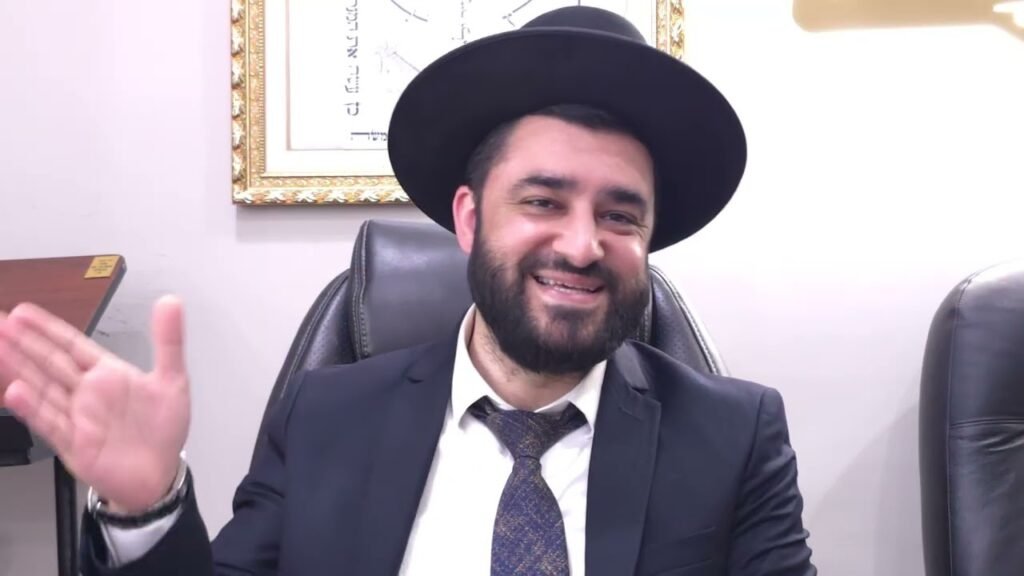Man in a dark suit and tie, wearing a wide black hat, smiling while waving his hand in an office setting with a framed picture on the wall behind him.