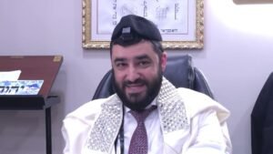 Bearded man wearing a kippah and silver-embroidered tallit, seated in an office with framed Hebrew artwork behind him, smiling at the camera.