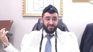 Bearded man in white ceremonial robe and kippah sits in an office chair, gesturing with his left hand as he speaks.