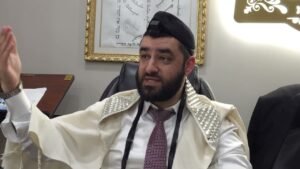 Bearded man wearing a kippah and prayer shawl sits at a desk, gesturing with his right hand during a discussion in an office setting, a framed artwork visible in the background.