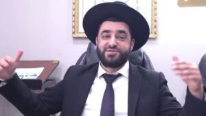 Man in a black suit and hat, gesturing with his hands in an office setting.