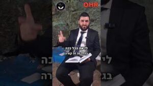 Bearded man in a suit sits outdoors against a rocky backdrop, reading from an open book while gesturing toward the camera; Hebrew captions appear on screen, with logos at the top corners.