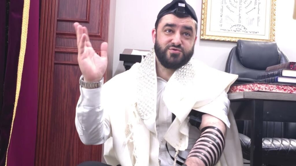 Bearded man wearing a white prayer shawl and tefillin, raising his right hand while speaking; tallit draped over his shoulders, tefillin straps on his left arm; office setting with books and a framed Hebrew script on the wall.