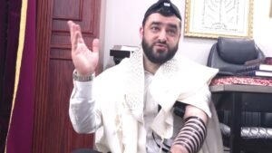 Bearded man wearing a white prayer shawl and tefillin, raising his right hand while speaking; tallit draped over his shoulders, tefillin straps on his left arm; office setting with books and a framed Hebrew script on the wall.