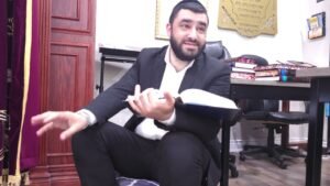 Man in a suit sits on the floor with an open book on his lap, gesturing with one hand in a study room.