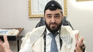 Bearded man wearing tefillin and a jeweled prayer shawl, seated at a desk, gesturing with his hands as he speaks.