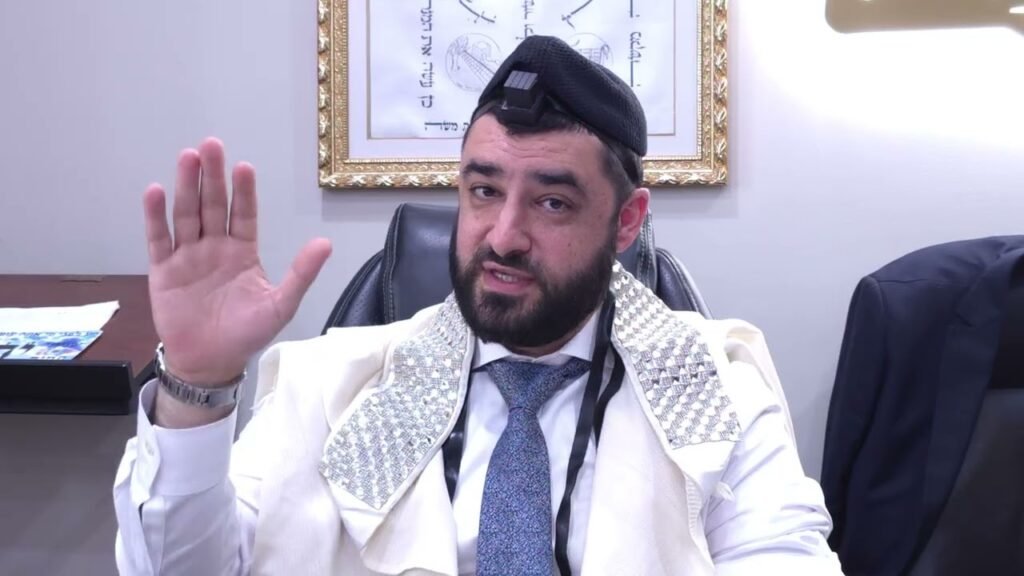 Bearded man in an office, wearing a white prayer shawl with silver studs and tefillin on his head, waving hello.