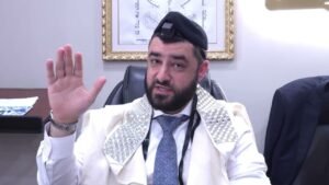 Bearded man in an office, wearing a white prayer shawl with silver studs and tefillin on his head, waving hello.