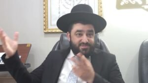 Bearded man in a black hat and suit gestures with his hands while speaking in an office setting.