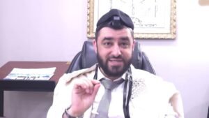 Bearded man wearing a kippah, tefillin on his head, a white prayer shawl over his shoulders, sitting in an office and gesturing with his hand.