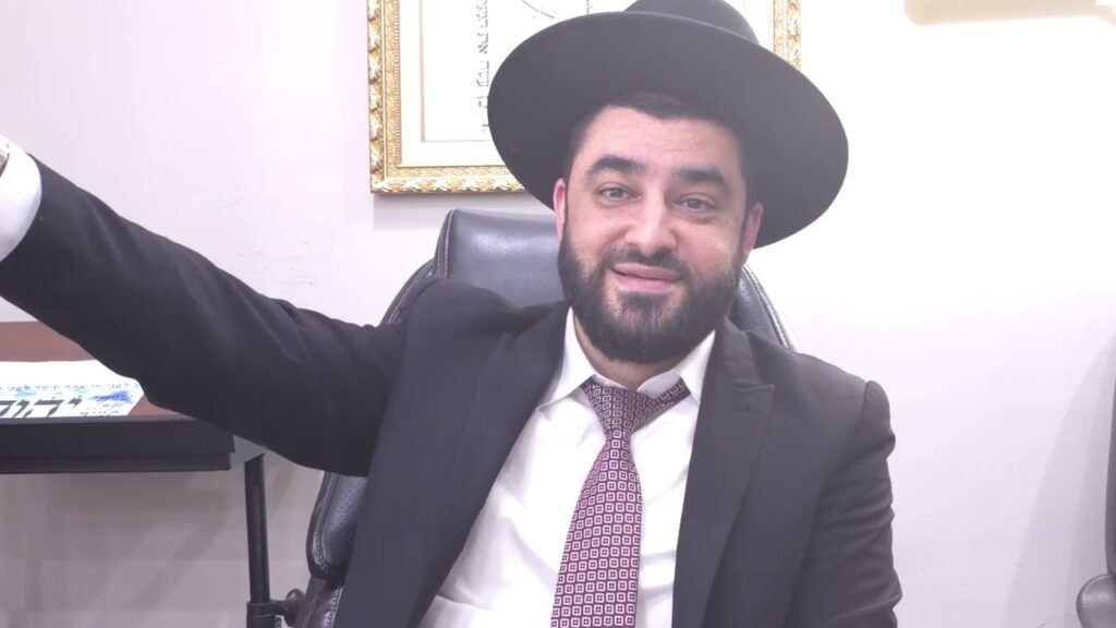 Bearded man in a black suit and hat sits at a desk, smiling with his left arm raised in a friendly gesture.