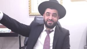 Bearded man in a black suit and hat sits at a desk, smiling with his left arm raised in a friendly gesture.