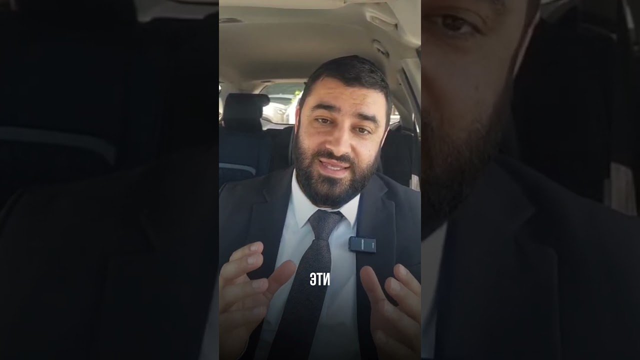 Man in a suit speaking to the camera from inside a car, with a clip-on microphone on his lapel and white subtitles on screen.