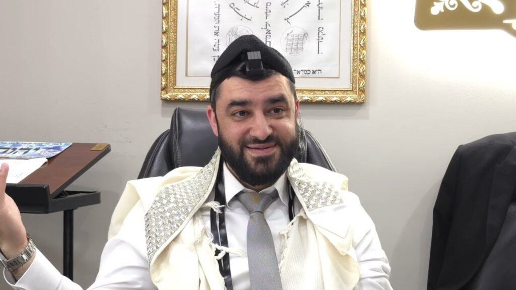 Bearded man in a white ceremonial shawl and tie, with a small camera mounted on his kippah, seated in an office with framed art behind him.