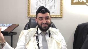 Bearded man in a white ceremonial shawl and tie, with a small camera mounted on his kippah, seated in an office with framed art behind him.