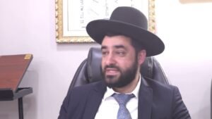 Bearded man in a suit and tie wearing a black hat seated in an office chair, framed artwork on the wall behind him.