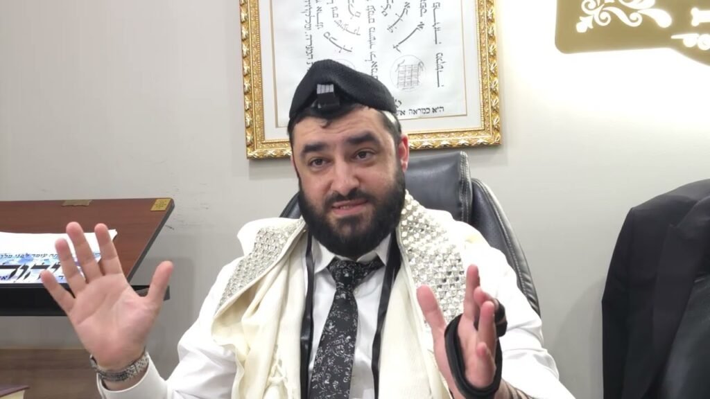 Bearded man wearing tefillin and tallit, seated at a desk, gesturing with hands during a talk in an office setting.