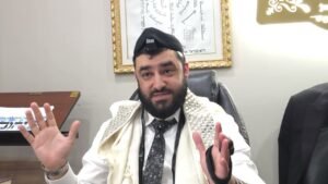 Bearded man wearing tefillin and tallit, seated at a desk, gesturing with hands during a talk in an office setting.