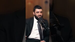 Man in a dark suit and tie speaks into a podcast microphone in a studio, wearing a kippah/fez and looking thoughtful
