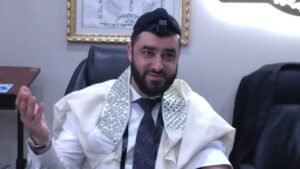 Bearded man in a white prayer shawl with sparkly sequin collars, wearing a kippah and tie, gesturing while seated in an office.