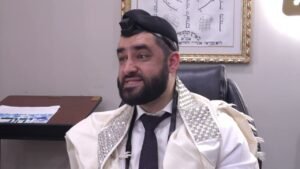 Man seated in an office, wearing a white tallit with silver embroidery and tefillin on his head, smiling slightly.