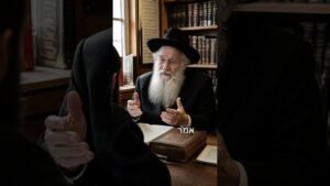 Elderly rabbi with a white beard and black hat speaks, gesturing with hands across a wooden desk piled with old books in a study.