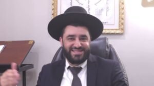 Smiling man with a beard in a dark suit and wide-brimmed black hat, seated in an office.