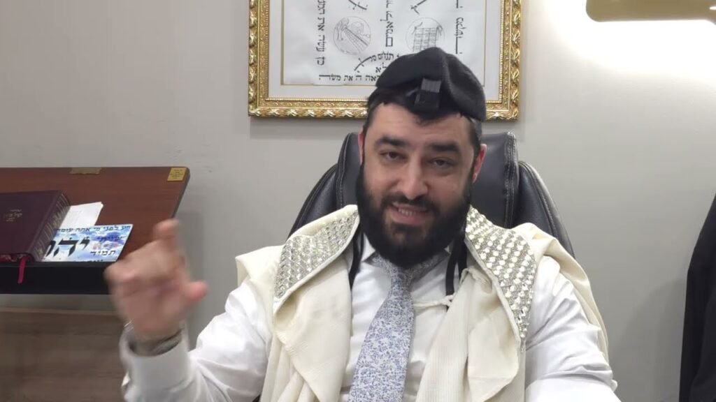 Bearded man wearing a kippah and tallit sits at a desk, gesturing with his hand while speaking.