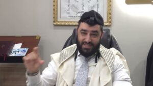 Bearded man wearing a kippah and tallit sits at a desk, gesturing with his hand while speaking.