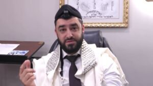 Bearded man in a white ceremonial robe with a jeweled collar sits in an office chair, gesturing toward the camera, with a framed artwork on the wall behind him and a desk to his left.