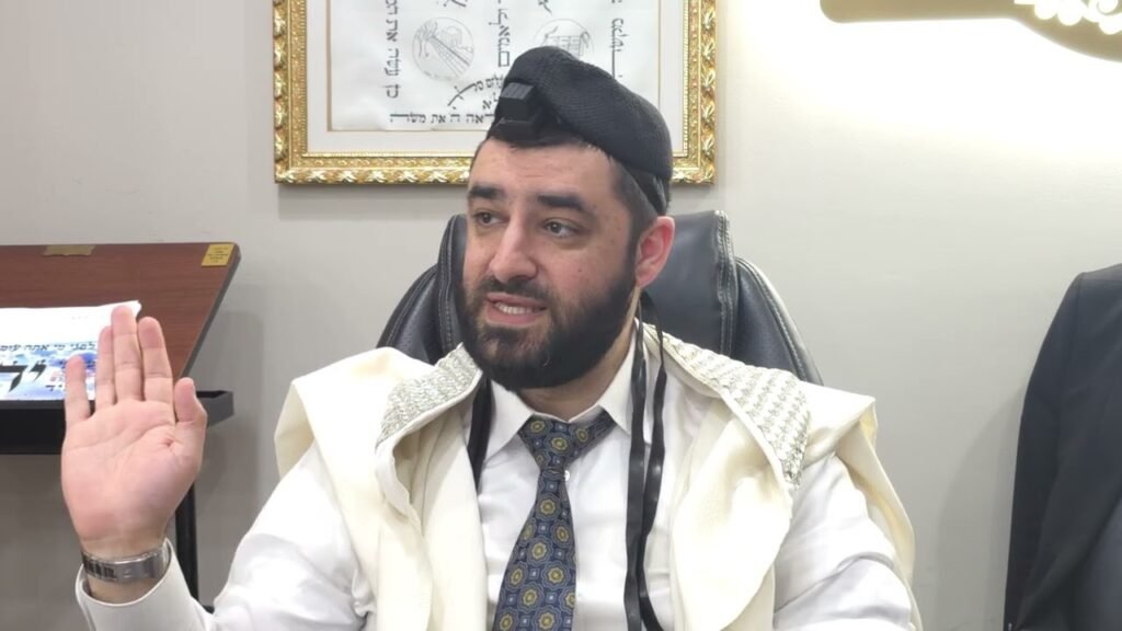 Bearded man in a white shirt, tie, kippah and prayer shawl raises his right hand while speaking in an office setting.