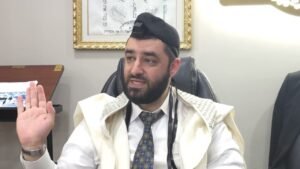 Bearded man in a white shirt, tie, kippah and prayer shawl raises his right hand while speaking in an office setting.
