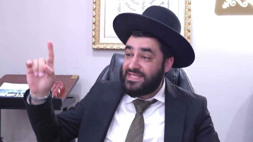 Bearded man in a black suit and hat sits at a desk, raising his index finger as if making a point in conversation.
