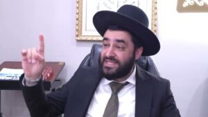 Bearded man in a black suit and hat sits at a desk, raising his index finger as if making a point in conversation.