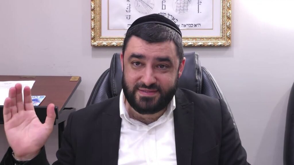 Bearded man wearing a kippah and suit sits at a desk in an office, raising his left hand to greet.