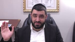 Bearded man wearing a kippah and suit sits at a desk in an office, raising his left hand to greet.