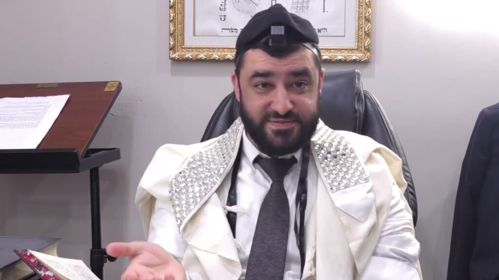 Bearded man wearing a white prayer shawl with silver-embellished shoulders and tefillin on his head, seated at a desk and speaking.
