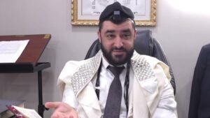 Bearded man wearing a white prayer shawl with silver-embellished shoulders and tefillin on his head, seated at a desk and speaking.