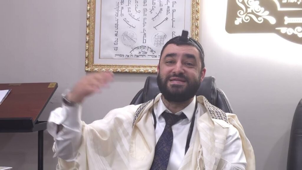 Man wearing a tallit and tefillin sits in a chair, gesturing with his hand during a discussion in an office setting framed by wall art behind him.