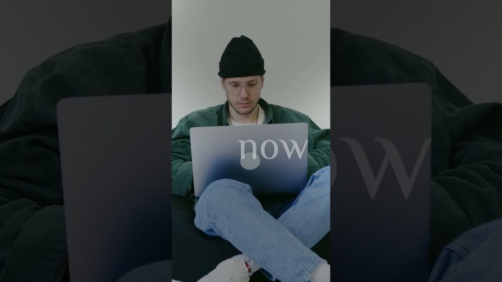 Man in a black beanie and glasses sits cross-legged on a dark surface, using a laptop with the word 'now' visible on the screen.