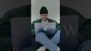 Man in a black beanie and glasses sits cross-legged on a dark surface, using a laptop with the word 'now' visible on the screen.