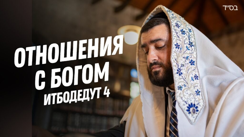 Bearded man wearing a white prayer shawl with blue embroidery, eyes closed in a prayerful moment inside a study or synagogue.