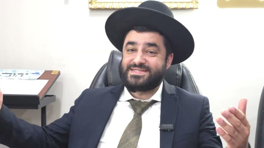 Smiling bearded man in a dark suit and wide-brimmed black hat seated in an office, gesturing with his hands.