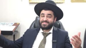 Smiling bearded man in a dark suit and wide-brimmed black hat seated in an office, gesturing with his hands.