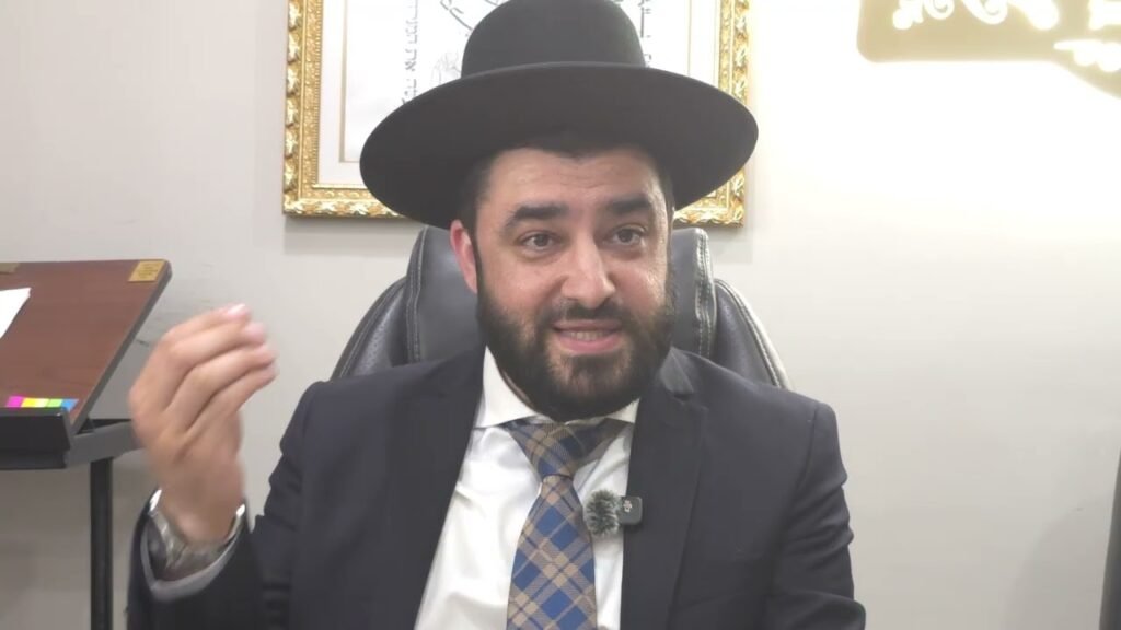 Bearded man in a black hat and suit, seated in an office, speaking into a lavalier microphone while gesturing with his hand.