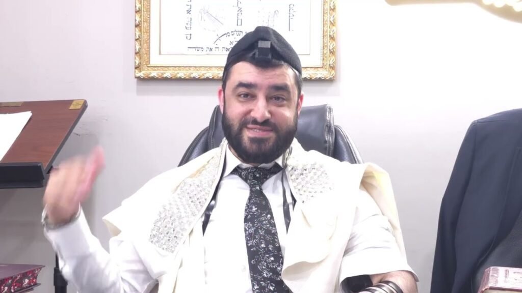 Bearded man in a white robe and kippah sits in an office chair, gesturing with his right hand, framed artwork on the wall behind him.
